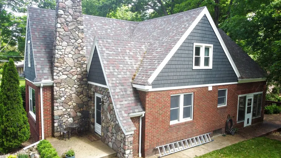 A house with a complex roofline features red brick and stone siding, gray shingles, and a tall stone chimney.